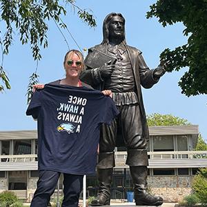 Kelly Scafariello holds a "Once a Hawk, always a Hawk" T-shirt by the Roger Williams statue on Bristol Campus
